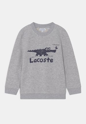 Sweat-shirt gris clair à manches longues avec poignets côtelés, présentant un crocodile noir caricatural et un oiseau avec le texte "Morning!" et le logo "Lacoste".