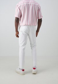 Kortärmad skär- och vitrandig skjorta i kombination med slim-fit vita byxor och vita sneakers med röda accenter. Neutral bakgrund.