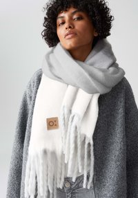 Grijze en witte sjaal gemaakt van zacht materiaal met franjes aan de uiteinden, voorzien van een tan kleurig label met een reliëf logo, gedragen over een grijze gestructureerde trui.