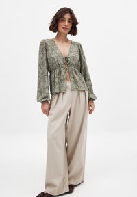 Blusa in pizzo floreale verde chiaro con scollatura a V, vita elastica e maniche lunghe a palloncino. Abbinata a pantaloni cream a gamba larga e scarpe marroni.