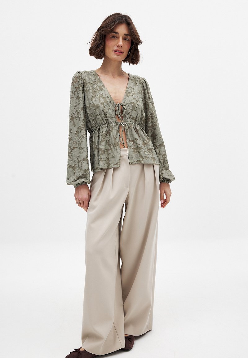 Blusa in pizzo floreale verde chiaro con scollatura a V, vita elastica e maniche lunghe a palloncino. Abbinata a pantaloni cream a gamba larga e scarpe marroni.