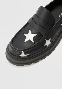 Svarta läderloafers med kraftig gummisula, utrustade med vita stjärnutskärningar och en rem över ovansidan. Slät textur och elegant design.