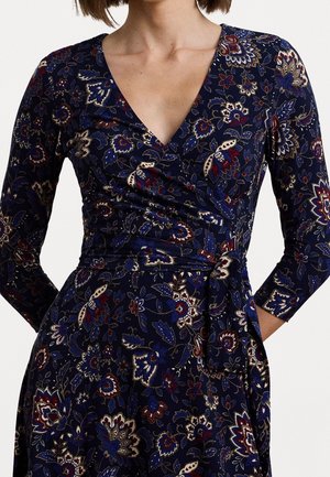 Femme portant une robe portefeuille fleurie bleu foncé avec des manches trois quarts et une ceinture nouée à la taille.