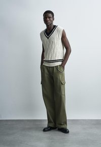 Mężczyzna ubrany w bezrękawnik w kolorze kremowym z czarnym wykończeniem, o splocie kablowym, oliwkowe spodnie cargo i czarne buty, stojący z rękami w kieszeniach.