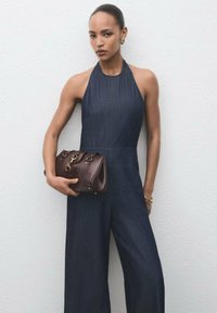 Denim jumpsuit med halterneck-design och vida ben, matchad med en brun läderhandväska med guldfärgade detaljer. Slät tygtextur.
