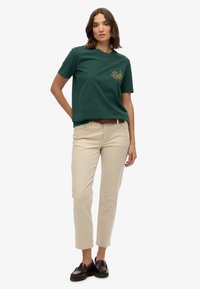 Donkergroen katoenen T-shirt met korte mouwen en een geborduurd ontwerp op de borst, gecombineerd met beige katoenen broek en donkere schoenen.