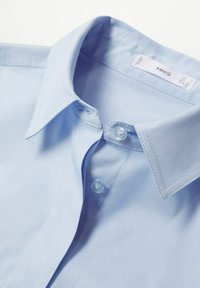 Chemise bleu clair à boutons, en coton lisse, avec un col classique, deux boutons et des coutures soignées le long des bords.