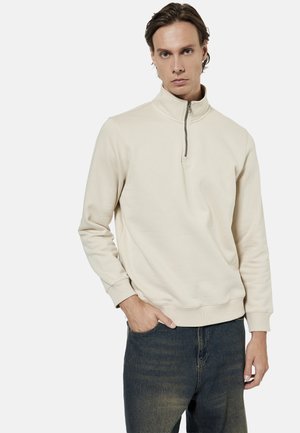 Ung mand iført en cremefarvet halv-zip trøje og mørkeblå jeans, med den ene hånd i lommen, mod en ensfarvet hvid baggrund.