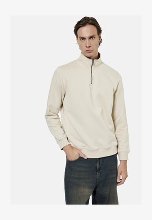 Ung mand iført en cremefarvet halv-zip trøje og mørkeblå jeans, med den ene hånd i lommen, mod en ensfarvet hvid baggrund.