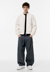 Light beige bomber jacket with a zip front, elastic cuffs, and two side pockets. Worn with loose, dark gray jeans and white sneakers.