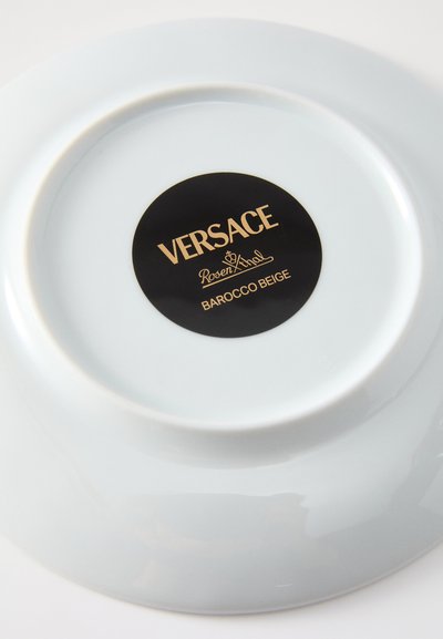 Porcelanowy talerz w jasnoniebieskim kolorze o gładkiej fakturze. Na talerzu znajduje się czarna i złota etykieta z napisem "Versace Barocco Beige." Okrągły kształt.