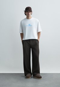 Wit katoenen T-shirt met blauw grafisch ontwerp, gecombineerd met donkerbruine wijde broeken en bruine instapschoenen met patroon, gedragen met een bruine pet.