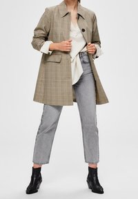 Kvinde iført en beige ternet frakke over en hvid skjorte, grå højtaljede jeans og sorte ankelstøvler, der står imod en ensfarvet lys baggrund.