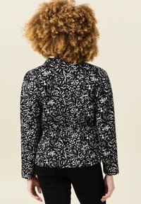 Chaqueta de tela negra con un patrón floral blanco, de manga larga, diseño ajustado y detalle en la cintura ceñida.