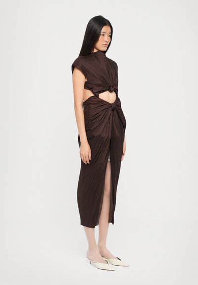 Robe plissée marron avec des découpes, des détails noués à la taille et aux épaules, et une fente haute. Le modèle porte des mules beiges et a de longs cheveux sombres.