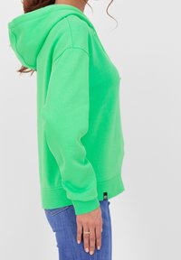 Donna che indossa una felpa con cappuccio verde brillante e jeans blu, in piedi di lato con la mano sinistra rilassata accanto a sé.