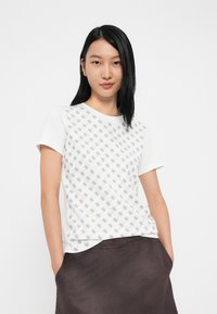 Hvit t-skjorte med korte ermer som har et gjentakende grått mønster. Klassisk design med rund hals og myk stoff, vist sammen med et mørkt skjørt.