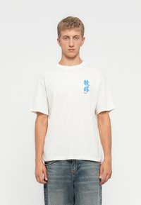 Young man facing forward wearing a white T-shirt with small blue graphic and text on the left chest, paired with faded blue jeans.
