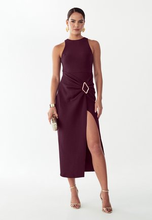Kvinde iført mørkelilla midikjole uden ærmer med høj slids og geometrisk spænde, der holder en clutch, guldringe og hæle.