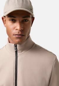Mand iført beige zip-up jakke og matchende kasket, neutral udtryk, vendt fremad mod en ensfarvet lys baggrund.
