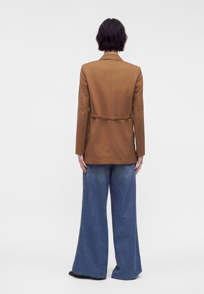 Personne aux cheveux courts et foncés portant un blazer marron et un jean bleu aux jambes larges, debout de dos sur un fond blanc uni.