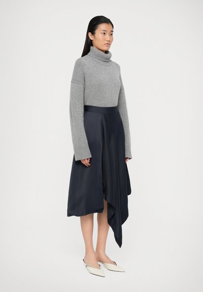 Maglione a collo alto grigio a maglia con maniche lunghe, abbinato a una gonna asimmetrica in seta blu scuro e scarpe a punta di colore chiaro.