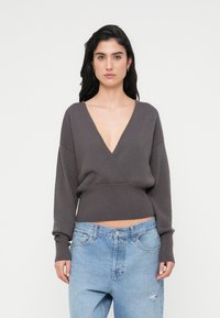 Σκούρο γκρι πλεκτό πουλόβερ με βαθύ V λαιμόκοψη και μακριά μανίκια, με κοντό τελείωμα. Συνδυάζεται με ανοιχτό μπλε τζιν με φθορές.