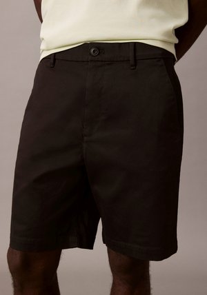 Czarne szorty o długości sięgającej połowy uda, z guzikiem i szlufkami na pasek, noszone przez osobę z jasnym topem wpuszczonym do środka.