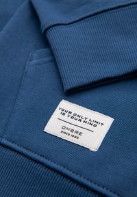 Niebieska bawełniana bluza ma Ribbed kołnierz oraz dół, z białą metką z napisem "TWÓJ JEDYNY LIMIT TO TWÓJ UMYSŁ" oraz "OMBRE od 1985".