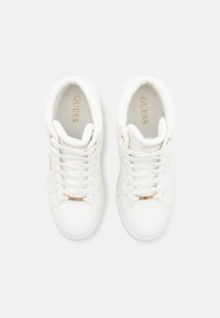 Baskets blanches en cuir lisse. Les caractéristiques incluent des lacets plats, des accents dorés et le nom de la marque "GUESS" sur les côtés et les semelles intérieures.