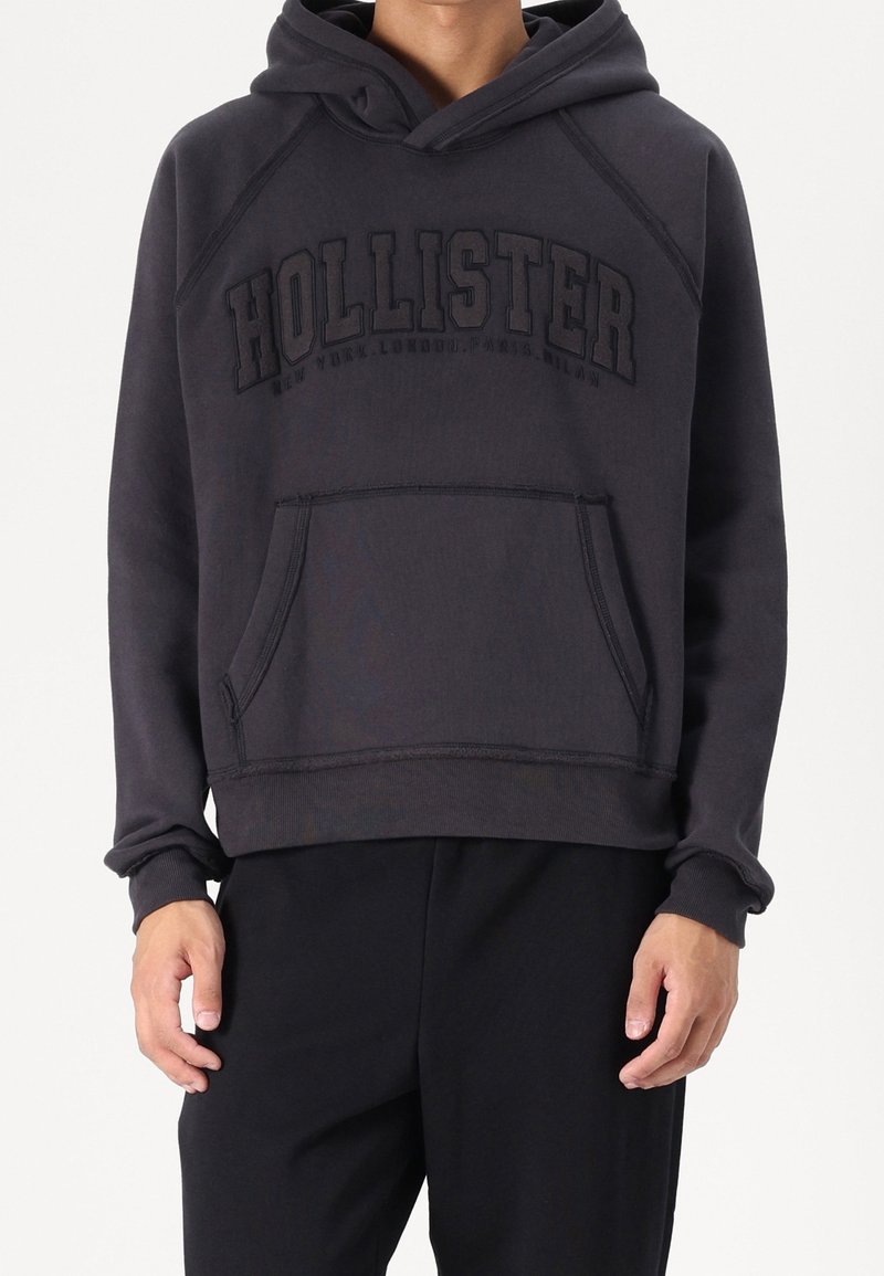 Personne portant un sweat à capuche Hollister gris foncé avec poche avant et capuche, assorti à un pantalon noir, sur un fond uni.