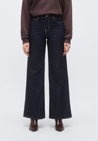 Sötétkék, széles szárú farmernadrág magas derékkal, öt zsebbel és kontrasztos varrással, barna bokacsizmával viselve.