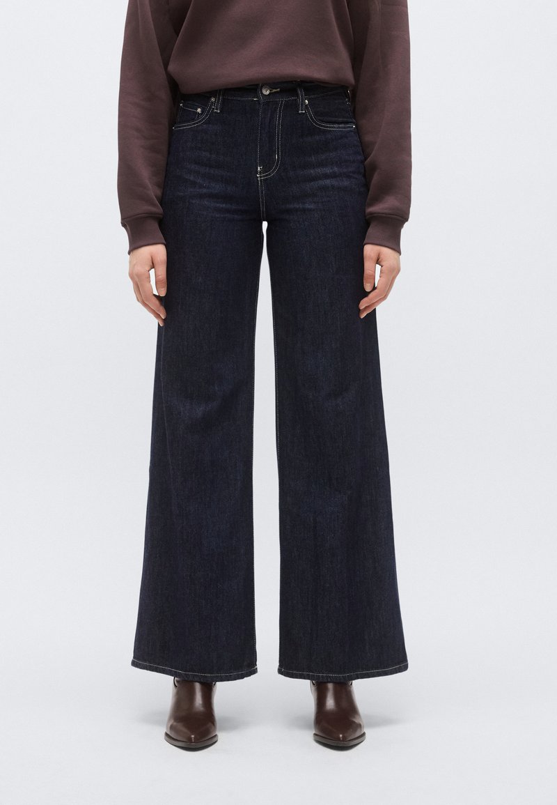 Sötétkék, széles szárú farmernadrág magas derékkal, öt zsebbel és kontrasztos varrással, barna bokacsizmával viselve.