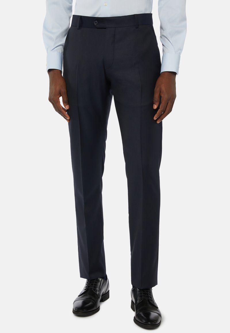 Man som bär marinblå kostymbyxor, svarta skinnskor och en ljusblå skjorta med lång ärm, stående mot en vit bakgrund.