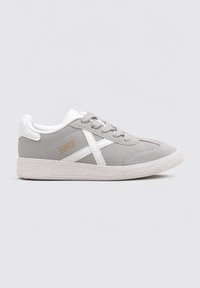 Zapatilla gris con suela blanca, que presenta un upper de tela texturizada, diseño de cordones y acentos blancos que incluyen un logo.