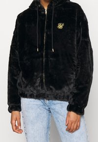 Sort faux fur zip-up hoodie med en guld-logo på brystet, har en hætte med snor og sidelommer. Blød tekstur, afslappet pasform.