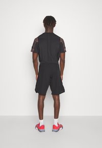 Sportoutfit mit einem schwarzen Kurzarmshirt mit strukturierten braunen Schultern und schwarzen Shorts, kombiniert mit roten und grauen Sportschuhen.