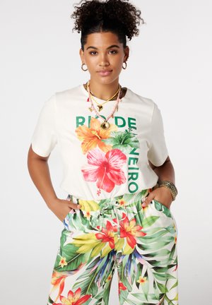 Vrouw met krullend haar, droeg hoepeloorbellen, gelaagde kettingen, een witte bloemige "Rio de Janeiro" T-shirt en kleurrijke tropische printbroek, handen in de zakken.
