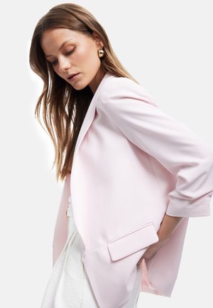 Frau mit langen braunen Haaren, die goldene Creolen, einen hellrosa Blazer und weiße Hose trägt, posiert mit der Hand in der Tasche.