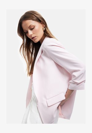 Frau mit langen braunen Haaren, die goldene Creolen, einen hellrosa Blazer und weiße Hose trägt, posiert mit der Hand in der Tasche.