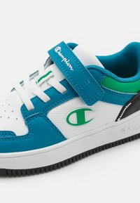 Blaue und weiße Sportschuh mit einem strukturierten Lederobermaterial, Schnürverschluss, grünen Akzenten und einer schwarzen Gummisohle. Champion-Logo an der Seite.