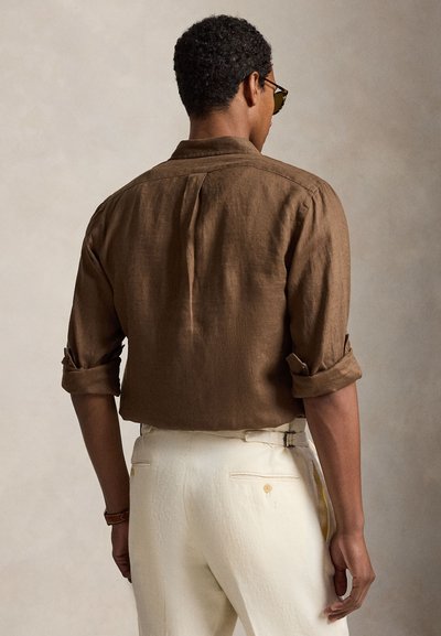 Bărbat cu mânecile suflecate, purtând o cămașă maro și pantaloni crem, stând cu spatele la un fundal neutru.