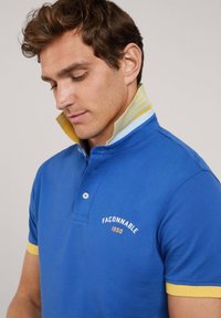 Niebieska koszulka polo z żółtymi akcentami. Posiada kołnierzyk zapinany na guziki i haftowane logo. Miękka bawełniana struktura, krótkie rękawy, dopasowany krój.