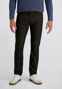 Uomo che indossa pantaloni slim-fit marrone scuro e sneakers bianche, in piedi contro uno sfondo chiaro e uniforme.