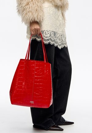 Osoba w beżowej puszystej kurtce i koronkowej bluzce, trzymająca dużą czerwoną torebkę o fakturze, ubrana w czarne spodnie i czarne buty na szpic.