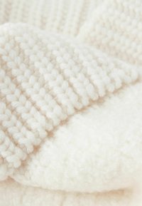 Kremowy, dziergany koc pokazujący teksturowane wzory; jedna strona ma gładkie, puszyste wykończenie, podczas gdy druga posiada żebrowany wzór.