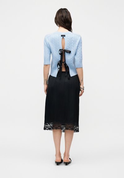 Femme vue de dos portant un haut en maille bleu clair à manches courtes avec des liens en ruban noir, une jupe midi noire ornée de dentelle et des escarpins noirs à talons bas.