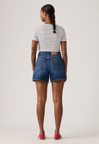 Γυναίκα που στέκεται με την πλάτη, φοράει κοντομάνικη ριγέ μπλούζα, σορτς από μπλε τζιν με ψηλή μέση, κόκκινα παπούτσια χωρίς τακούνι, με πλεγμένα μαλλιά.