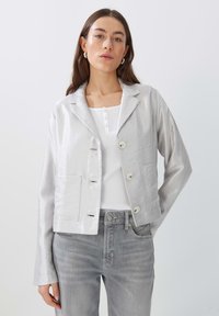 Silberfarbene, strukturierte, kurz geschnittene Jacke mit Reverskragen, einreihigen Knöpfen und zwei vorderen Taschen, kombiniert mit einem weißen Oberteil und grauen Jeans.