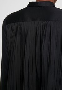 Schwarze Jacke mit einem gefalteten Rückendesign, glatter Stofftextur und klassischem Kragen. Die Falten erzeugen vertikale Linien für zusätzliche Details.